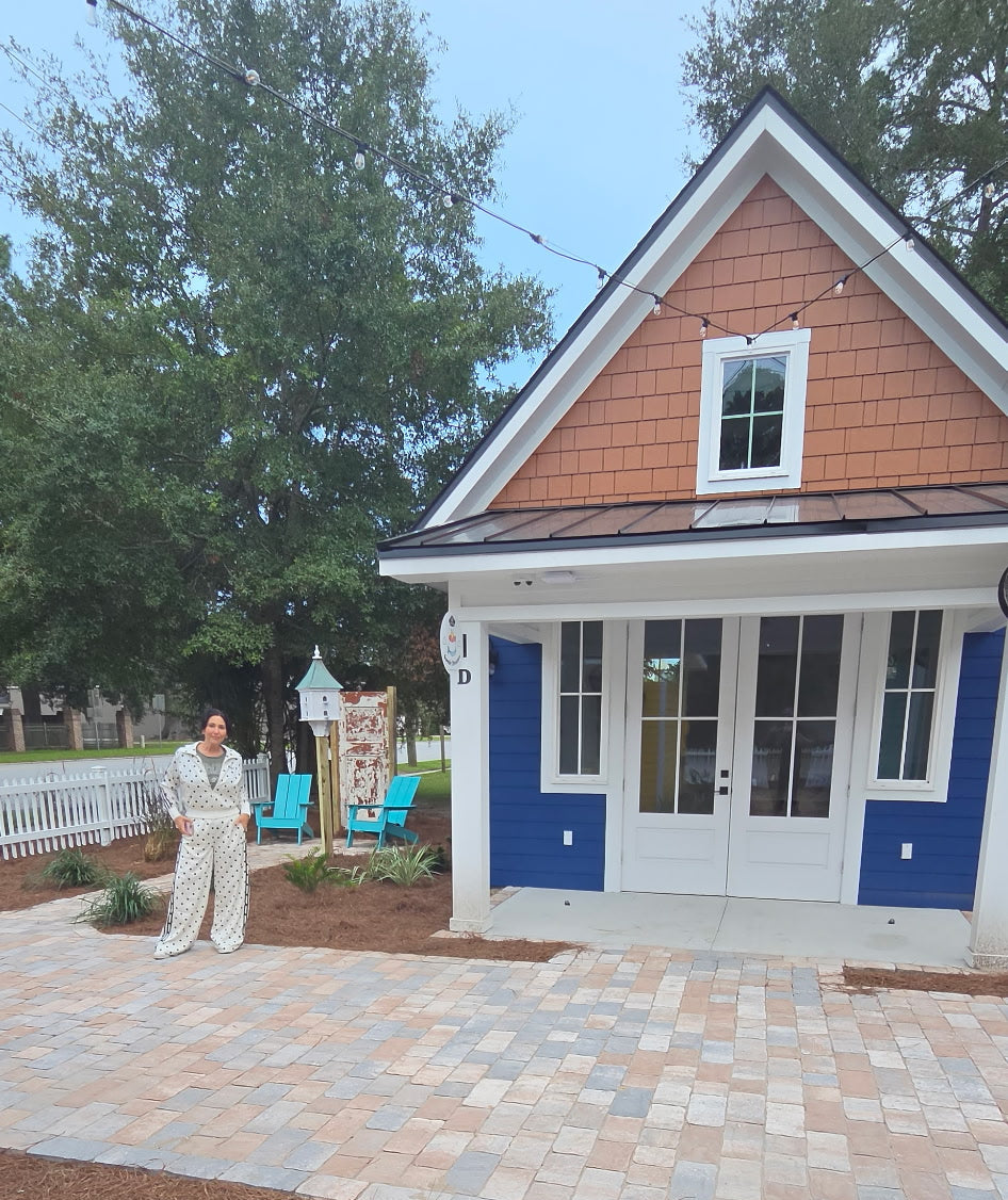 A person in a light beige outfit stands on a newly installed paver patio in front of a small house with blue shutters, white trim, and a brown shingled gable roof. Mulch covers the foreground, with trees and a yard visible behind.