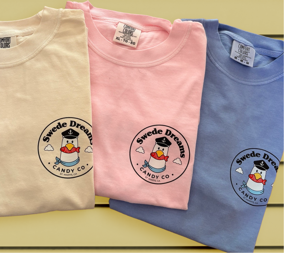 Four folded short-sleeve T-shirts in white, cream, pink, and blue, each printed with a circular 'Sweede Dream Candy Co.' logo featuring a penguin in a sailor hat.
