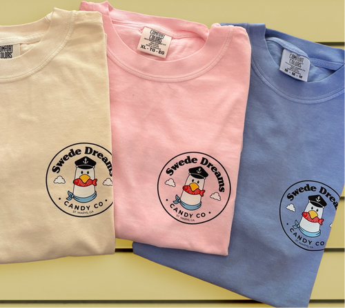 Four folded short-sleeve T-shirts in white, cream, pink, and blue, each printed with a circular 'Sweede Dream Candy Co.' logo featuring a penguin in a sailor hat.